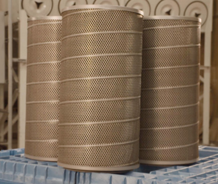 Three replacement cartridge filters stacked on a flat surface and positioned in front of a dust collector system in an industrial factory.